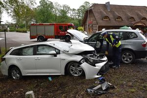 Miejsce wypadku. Wrak białego samochodu. Policjant przykłada do niego taśmę mierniczą. W tle srebrny samochód z podniesioną pokrywa silnika oraz wóz bojowy straży pożarnej.