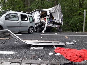 Na jezdni leżą elementy z rozbitych pojazdów oraz płachta czerwonego materiału. W tle srebrny samochód osobowy oraz uszkodzony tył ciężarówki.