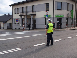 Policjant ruchu drogowego stoi na jezdni wstrzymując ruch pojazdów. Przed nim wybiegający z drogi podporządkowanej uczestnik zawodów.