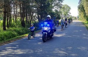 Policjant na motocyklu. Włączone sygnały świetlne. Obok niego dziecko na małym rowerku w kasku i kamizelce odblaskowej. Za nimi sznur rowerzystów.