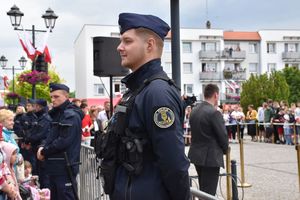 Policjanci w umundurowaniu ćwiczebnym stoją w kordonie przed metalowym ogrodzeniem, za nimi teren wyznaczony widoczny mężczyzna z słuchawką w uchu. W tle widoczne zabudowania oleskiego rynku.