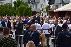 Pan Prezydent stoi pośród zgromadzonych na oleskim rynku mieszkańców. Na pierwszym planie w lewym dolnym rogu widoczny policjant, który stoi przed metalowym ogrodzeniem.