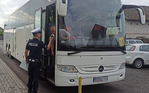 Policjant sprawdza stan trzeźwości kierowcy, który stoi na schodkach autokaru w otwartych drzwiach. Kierowca w dmucha w urządzenie pomiarowe.