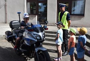 Policyjny motocykl. Przy nim stoi policjant ruchu drogowego w czapce i zielonej odblaskowej kamizelce. Na motocyklu siedzi mały chłopiec, a obok stoi troje innych dzieci.