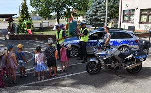 Policyjny motocykl, radiowóz. Obok nich stoi dwóch policjantów w czapkach na głowie i żółtych kamizelkach odblaskowych. Przed nimi po lewej stronie stoi grupka małych dzieci.