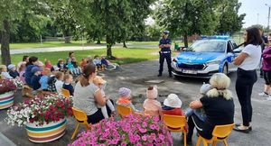 Na pierwszym planie dzieci i opiekunki siedzące na krzesełeczkach. Przed nimi radiowóz stojący przodem a obok niego po lewej stronie policjant ruchu drogowego.