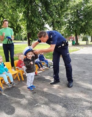 Małe dzieci siedzą w szeregu w fotelikach. Jedno dziecko stoi przed policjantem, który zakłada mu na głowę policyjną czapkę.