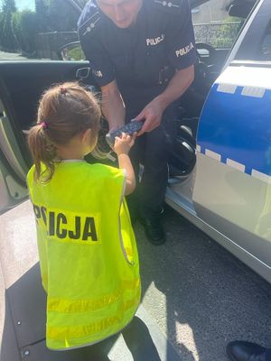 Mała dziewczynka w żółtej kamizelce odblaskowej z napisem POLICJA stoi przy otwartych drzwiach radiowozu. Obok niej policjant, który trzyma w rękach pilot od włączania świateł i sygnałów uprzywilejowania. Dziewczynka naciska klawisze na pilocie.