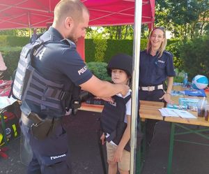 Policjant ubrany w kamizelkę taktyczną pomaga zapiąć hełm ochronny stojącemu przed nim małemu chłopcu. Chłopiec ma założoną kamizelkę kuloodporną z napisem na piersi POLICJA, a w prawej ręce trzyma pałkę wielofunkcyjną typu TONFA.