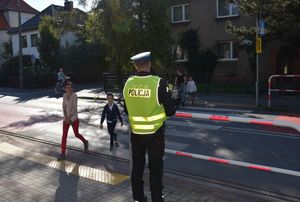 Przejście dla pieszych. Policjant stoi na chodniku i obserwuje drogę. Na przejściu piesi.