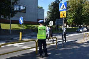 Przejście dla pieszych. Policjant stoi przy przejściu i obserwuje ruch na drodze. Na przejściu trzech pieszych.