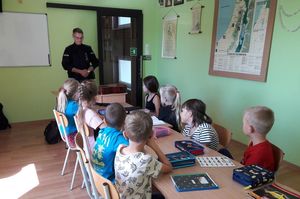 klasa lekcyjna, dzieci siedzą przy stole, patrzą w stronę tablicy, na środku stoi policjant.