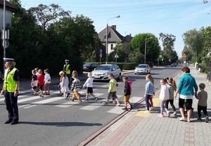 grupa przedszkolaków przechodzi po pasach  w asyście dwóch policjantek ruchu drogowego. W tle na jezdni stojące samochody.