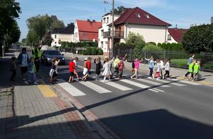 grupa przedszkolaków przechodzi po pasach z rękami uniesionymi w górę, policjantka stoi na jezdni z uniesioną ręką, za nią stoi samochód.