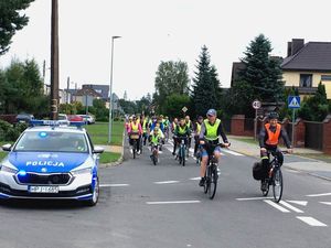 Peleton przejeżdża przez skrzyżowanie, obok stoi radiowóz na sygnałach. W tle widoczne zabudowania.