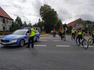 Policjant stoi przy radiowozie na środku skrzyżowania, obok przejeżdżają uczestnicy rajdu. W tle zabudowania i niebo.