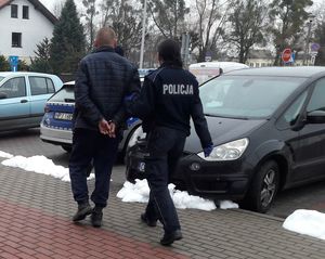 Policjantka prowadzi mężczyznę z założonymi kajdankami na ręce trzymane z tyłu. W tle parking z zaparkowanymi pojazdami.