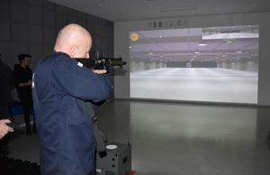 Komendant Powiatowy Policji w Oleśnie w postawie strzeleckiej stojąc celuje w wyświetlaną na ścianie tarczę strzelecką. W rękach trzyma replikę karabinka M4.