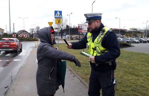 Okolica przejścia dla pieszych. Policjant ruchu drogowego zakłada odblaskowa opaskę na przedramię kobiecie.