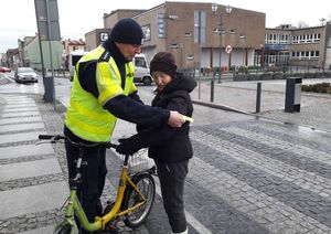 Plac Wolności w Dobrodzieniu. Policjant w odblaskowej kamizelce zakłada odblaskową opaskę na ramię kobiety prowadzącej rower w rejonie przejścia dla pieszych.