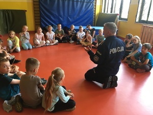 Policjant w klęku na parkiecie sali gimnastycznej, do koła niego siedzą dzieci.