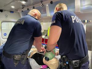 Wnętrze karetki. Dwaj policjanci prowadzą na fantomie resuscytację krążeniowo-oddechową przy pomocy worka samorozprężalnego.