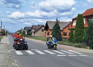 Ulica w Dobrodzieniu. Policjant na motocyklu i dwaj strażacy na quadzie zabezpieczają trasę biegu.