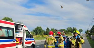 Miejsce wypadku drogowego. Po prawej stronie stoi karetka pogotowia ratunkowego. W tle grupa strażaków nad nimi lądujący śmigłowiec LPR.