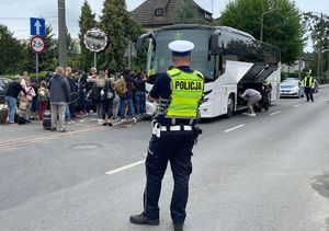 Ulica Krasickiego w Oleśnie, okolica Domu Harcerza. Policjant ruchu drogowego wstrzymuje ruch pojazdów. W tle autokar i uczestnicy wyjazdu z bagażami.