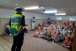 Umundurowany policjant ruchu drogowego stoi przed dziećmi siedzącymi przed nim na podłodze. Za plecami trzyma małe koło w kolorze zielonym.