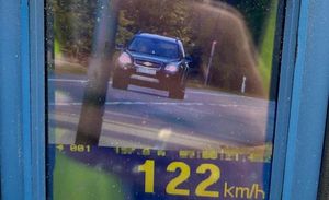 Urządzenie pomiarowe do mierzenia prędkości. Na wyświetlaczu żółte cyfry 122km/h. Nad nimi zdjęcie ciemnego samochodu osobowego.