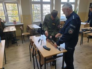 Policjant instruuje ucznia podczas rozkładania broni - karabinku AKMS. Elementy broni są rozłożone na stoliku. W tle okna.