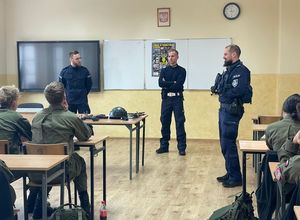 uczniowie w zielonych mundurach siedzą w ławkach szkolnych, przed nimi - przy tablicy stoją trzej policjanci. Na tablicy przyklejony jest plakat o tematyce promującej służbę zatytułowany : &quot; zawsze w pierwszym szeregu&quot;.