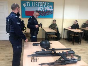 Na pierwszym planie stolik z bronią, za stolikiem stojący bokiem dwaj policjanci, jeden z nich trzyma w rękach broń służbową krótką. W tle w ławkach szkolnych siedzą uczniowie w zielonych mundurach, na ścianie plakat z napisem 11 listopada i biało-czerwoną flagą.