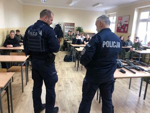 Na pierwszym odwróceni  plecami dwaj policjanci, w tle w ławkach szkolnych siedzą uczniowie w zielonych mundurach.