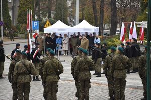 Żołnierze stoją w dwuszeregu plecami. W tle widoczni zaproszeni goście, którzy stoją pod białym zadaszeniem - namiotem. Po prawej stronie biało czerwone flagi.