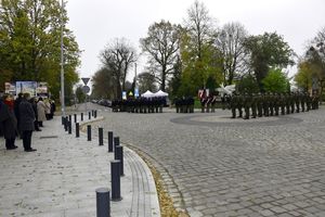 Fragment ulicy i chodnika, od lewej  na chodniku stoją uczniowie i nauczyciele. Po prawej stronie stoją uczniowie w mundurach ustawieni w szeregach wraz z pocztem i sztandarem szkoły, obok nich stoją Żołnierze Wojska Polskiego ustawieni w szeregach. Pomiędzy stojącymi mundurowymi Pomnik Powrotu Ziem Piastowskich do Macierzy . W oddali cmentarz miejski.