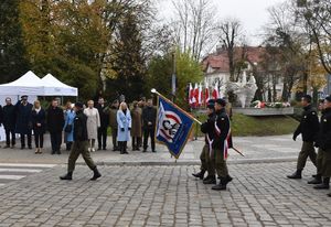 Po ulicy maszerują uczniowie w mundurach ze sztandarem szkoły. Za nimi Zaproszeni goście w tym przedstawiciele Wojska Polskiego, Policji, Straży Pożarnej i Nadleśnictwa, którzy  trzymają prawe dłonie przy głowie na gest oddania honoru. W tle Pomnik Powrotu Ziem Piastowskich do Macierzy.