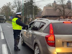 Policjant stoi na jezdni przy pojeździe osobowym. ma wyciągniętą dłoń w stronę kierowcy, w której trzyma urządzenie do pomiaru stanu trzeźwości. W tle wyjeżdżający z drogi prostopadłej pojazd ciężarowy.