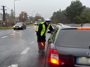 Policjant stoi na jezdni przy pojeździe osobowym. ma wyciągniętą dłoń w stronę kierowcy, w której trzyma urządzenie do pomiaru stanu trzeźwości. w drugiej dłoni trzyma latarkę z czerwoną nakładką. W tle sznur samochodów.