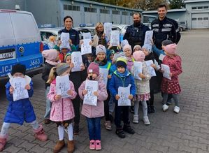 przedszkolaki stoją w grupie przed policjantem obok radiowozu, trzymają w rękach kartki do kolorowania z wizerunkiem psa z kreskówki &quot; psi patrol&quot;.