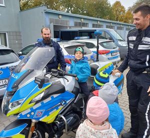 przedszkolak siedzi na policyjnym motocyklu obok policjant w kombinezonie motocyklowym. obok motocykla stoi kilku przedszkolaków, w tle stoi policjant w umundurowaniu służbowym.