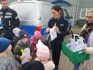 Policjantka wręcza dzieciom elementy odblaskowe. Obok stoi policjant w kombinezonie motocyklowym na tle radiowozu.