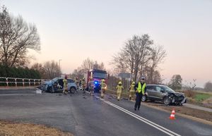 Na jezdni - skrzyżowaniu dwa rozbite pojazdy osobowe, w tle pojazd Państwowej Straży Pożarnej. Na pierwszym planie policjant a tuż za nim Strażacy.