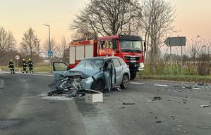 NA jezdni rozbity pojazd osobowy, tuż za nim wóz Państwowej Straży Pożarnej. Obok po lewej stronie Strażacy.