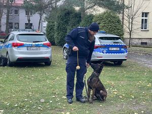 policjant  w ręku trzyma piłeczkę na którą spogląda siedzący obok nogi  pies.