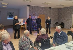 Na pierwszym planie siedzący przy stole seniorzy, tuz za nimi stojący na środku dwaj policjanci i policjantka oraz Pani rzecznik konsumentów. W tle stojąca choinka.