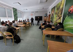 Zdjęcie wykonane w kolorze, przedstawia uczniów siedzących w szkolnych ławkach, po prawej stronie na ścianie fototapety z wizerunkami owadów: biedronka i ważka. Na końcu klasy w środkowej części kadru stoją policjant i policjantka. Po lewej stronie okna częściowo zasłonięte  roletami zewnętrznymi.