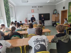 Zdjęcie wykonane w kolorze, przedstawia uczniów siedzących w szkolnych ławkach,  Na końcu klasy w środkowej części kadru stoją policjant i policjantka. Po lewej stronie okna częściowo zasłonięte  roletami zewnętrznymi.