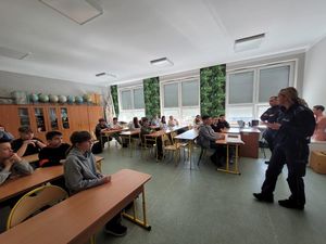 Zdjęcie wykonane w kolorze, przedstawia od lewej strony  uczniów siedzących w szkolnych ławkach, za uczniami meblościanka, na której ustawione jest obok siebie kilka kolorowych globusów oraz inne przybory szkolne , po prawej stronie stoją policjantka i policjant.Za nimi  okna częściowo zasłonięte  roletami zewnętrznymi.